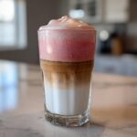 A tall glass showcasing the distinct layers of a strawberry cold foam latte: milk, espresso, and pink foam.